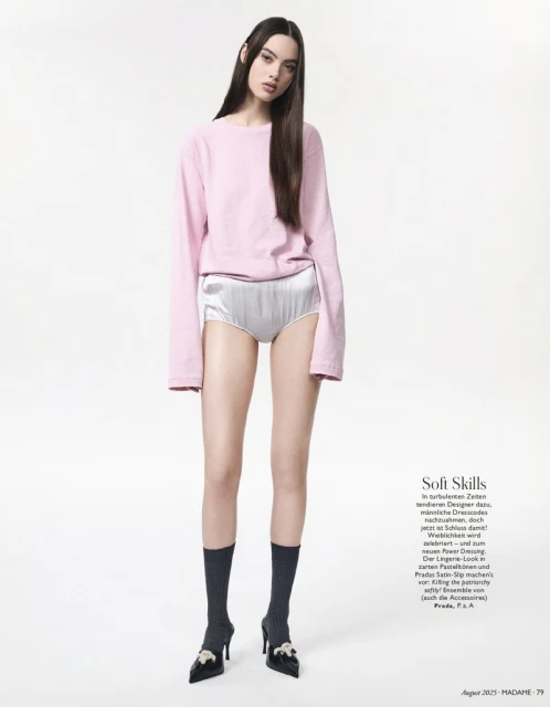 Full-length shot of a young woman in a pink sweater and silver shorts, styled with dark socks and high heels against a white backdrop.
