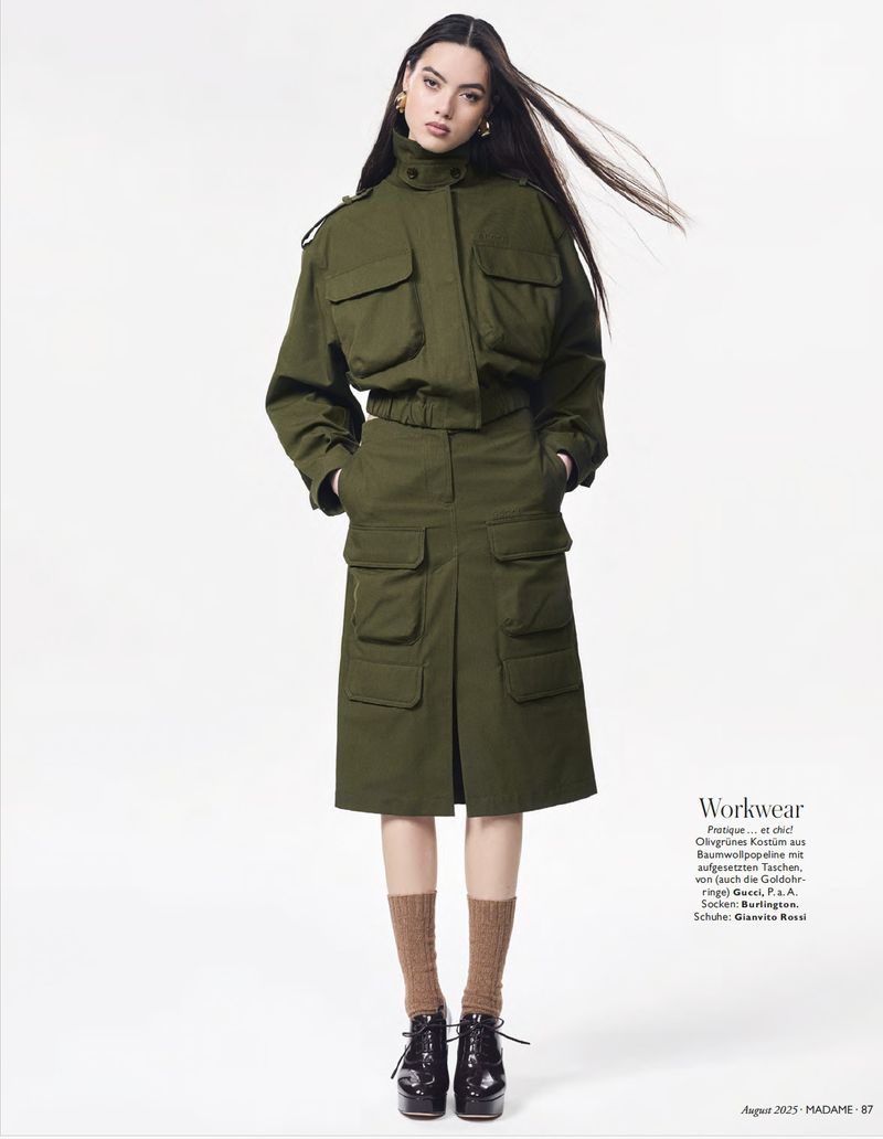 A woman in an olive green utility jacket and skirt stands in a studio with long dark hair, brown socks, and patent leather shoes.