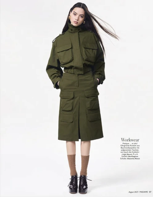 A woman in an olive green utility jacket and skirt stands in a studio with long dark hair, brown socks, and patent leather shoes.