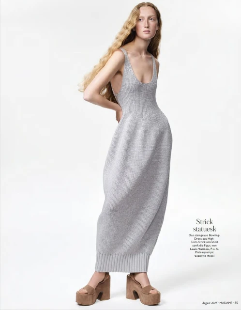 A fair-skinned woman with long blonde hair poses in a light grey knit dress and brown platform sandals against a white backdrop.