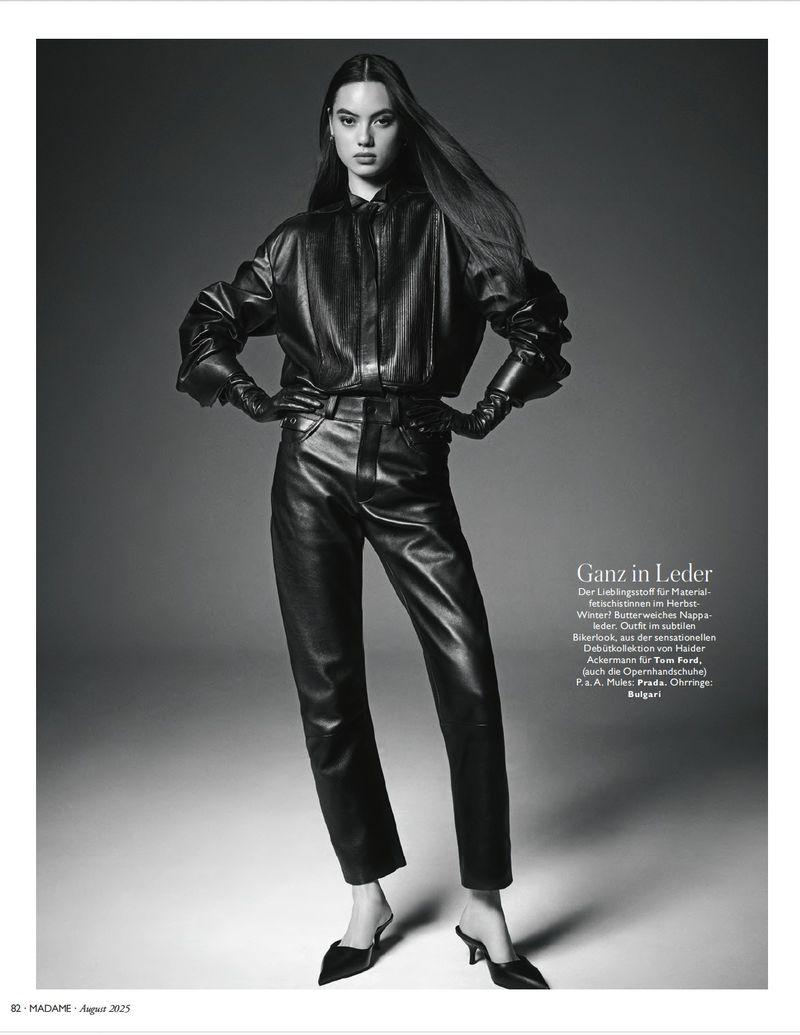 Monochrome fashion shot of a slender woman in a black leather outfit with mules, captured in a full shot against a plain gray backdrop.