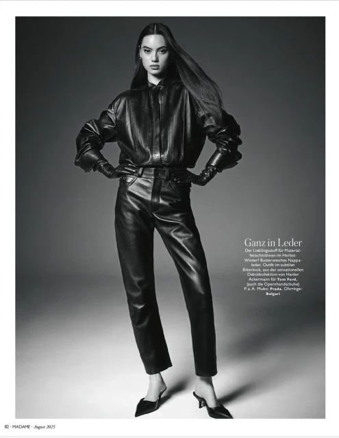 Monochrome fashion shot of a slender woman in a black leather outfit with mules, captured in a full shot against a plain gray backdrop.