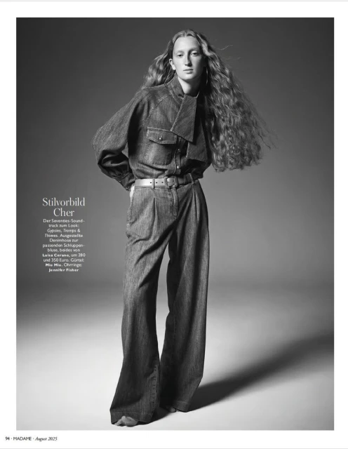 Fashion editorial shot: a stylish young woman stands in wide-leg denim suit with long, wavy hair against a neutral gradient backdrop.