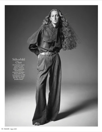 Fashion editorial shot: a stylish young woman stands in wide-leg denim suit with long, wavy hair against a neutral gradient backdrop.