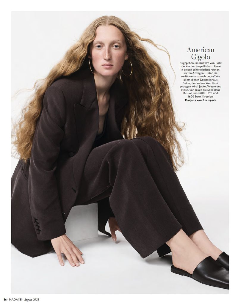 A woman with long wavy hair poses in a dark blazer and wide-legged trousers, seated against a white backdrop in a studio setting.