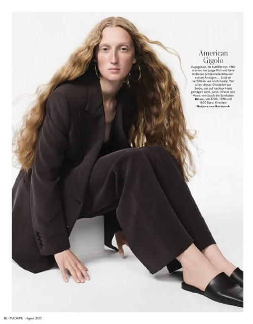 A woman with long wavy hair poses in a dark blazer and wide-legged trousers, seated against a white backdrop in a studio setting.