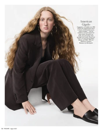 A woman with long wavy hair poses in a dark blazer and wide-legged trousers, seated against a white backdrop in a studio setting.