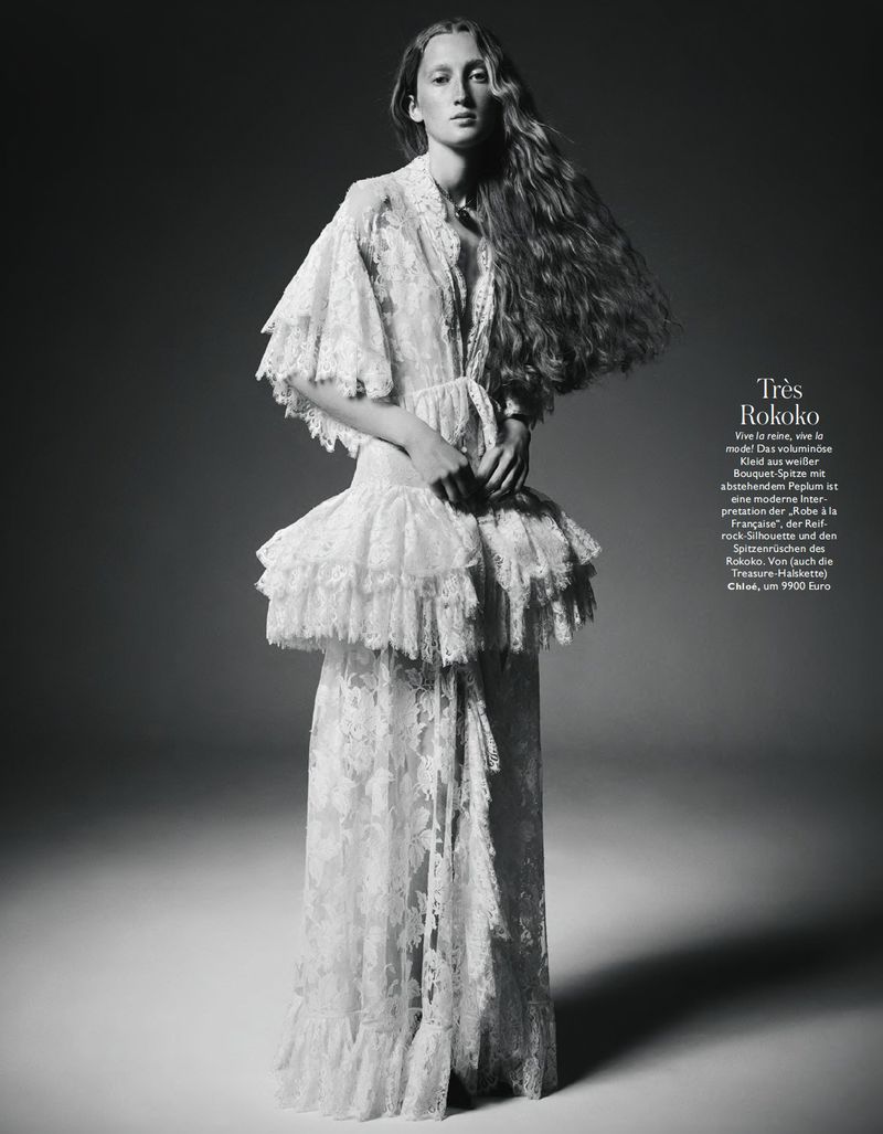 Model with long textured hair posing in an ornate tiered white lace gown against a smooth studio background.