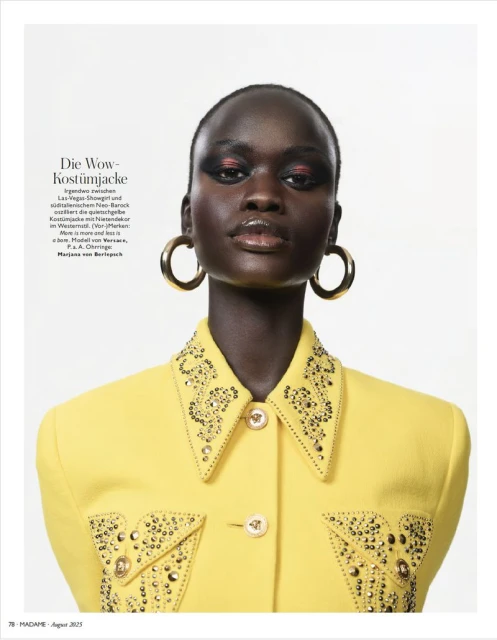 Headshot of a dark-skinned fashion model wearing a yellow jacket with gold embellishments and large gold hoop earrings on a white background.