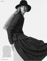 Fashion model wearing a black wide-brim fedora and a textured dark tweed jacket with matching skirt against a white studio background.