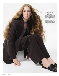 Model with long wavy hair wearing a dark brown tailored suit and black mules, seated on a white block against a white studio background.