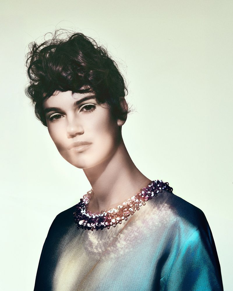 Model with short dark curls wearing an iridescent blue top with a beaded collar against a pale seamless background.