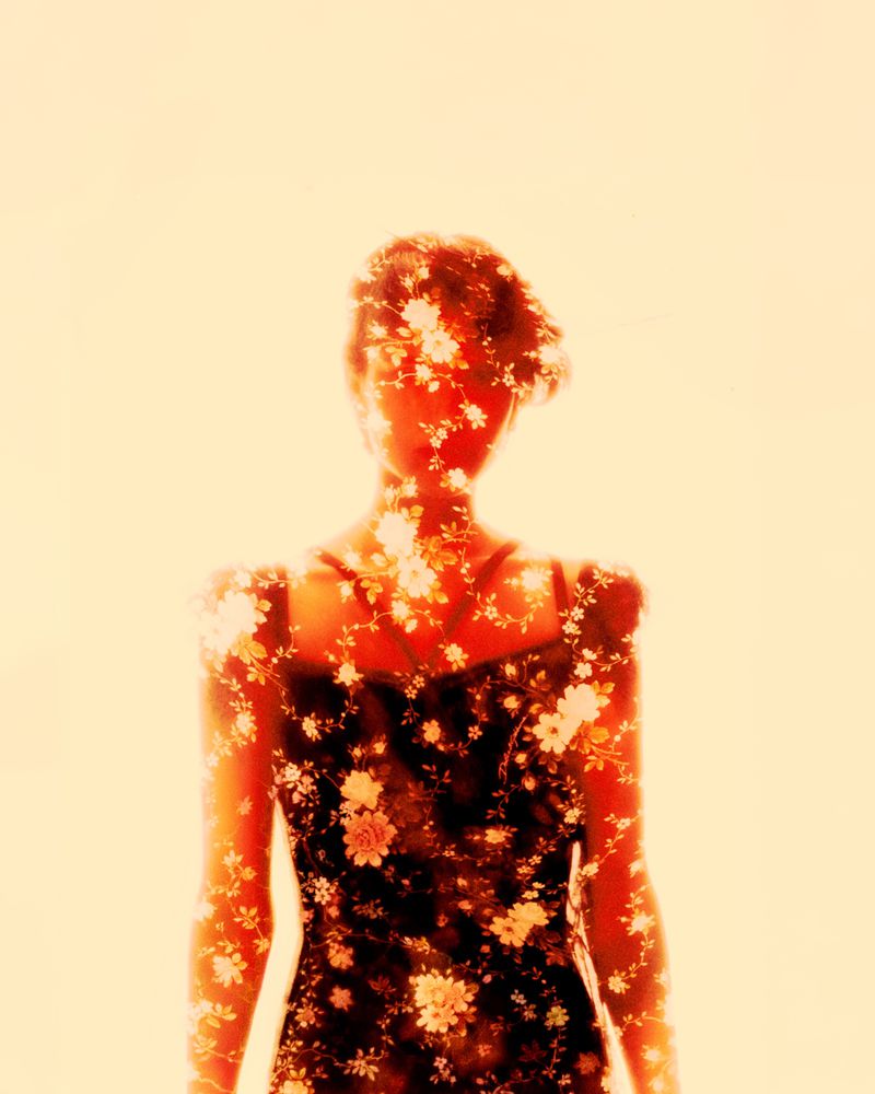 Double exposure portrait of a person in a floral dress, overlaid with delicate white flower patterns against a soft warm light background.