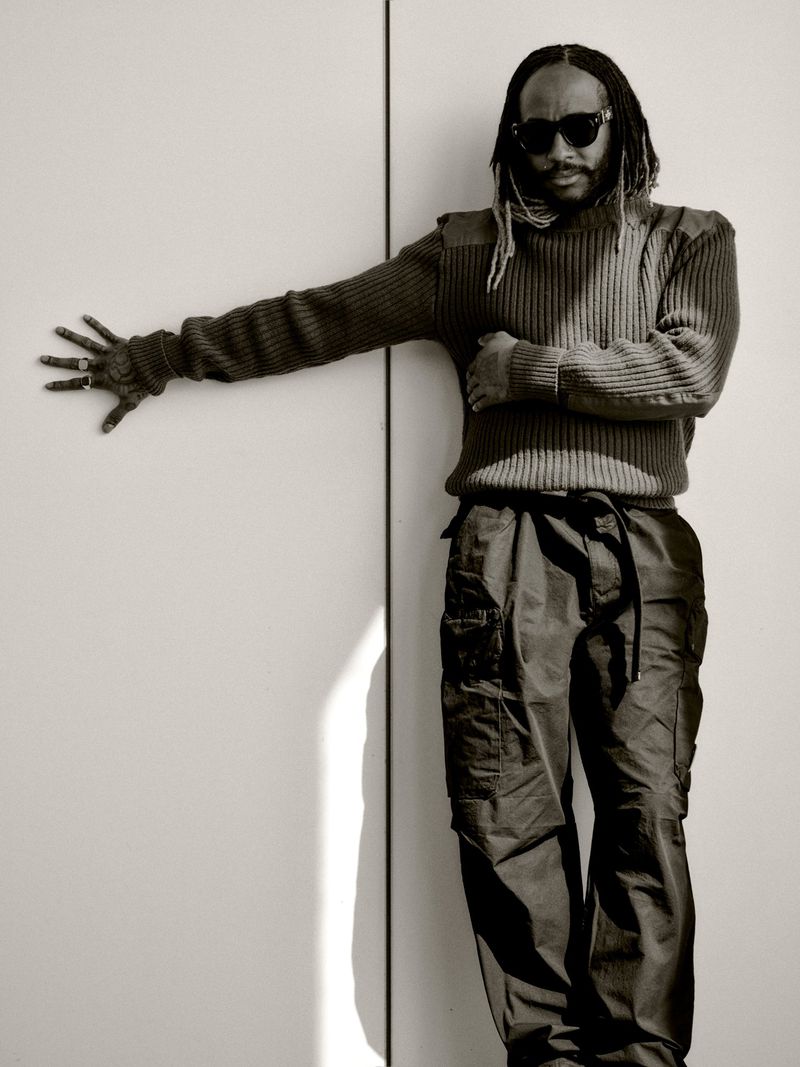 Thundercat wearing sunglasses, a ribbed knit sweater, and cargo pants, posing against a divided neutral wall.
