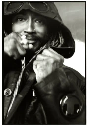 Black and white portrait of Thundercat wearing a hooded jacket with goggles on the hood, gripping the collar.