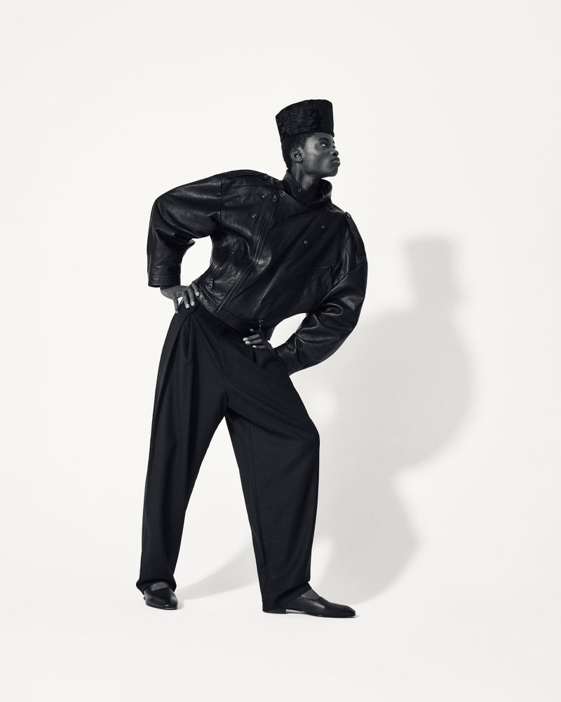 A Black male model posing in an Edward Crutchley oversized black leather jacket and wide-leg black trousers against a white background.
