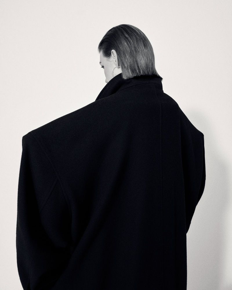 Model seen from behind wearing an exaggerated oversized black sculptural coat against a white studio background.