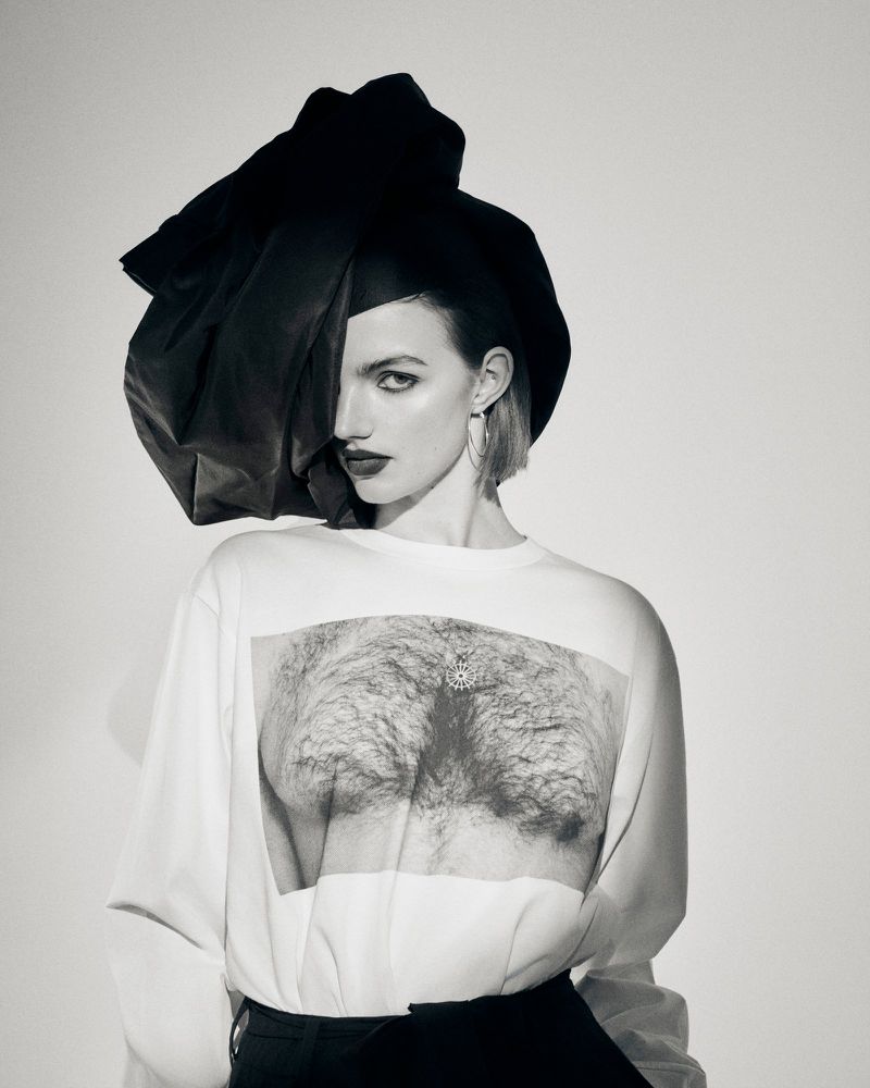 Model wearing an exaggerated black headpiece and a white printed long-sleeve top against a light studio backdrop.