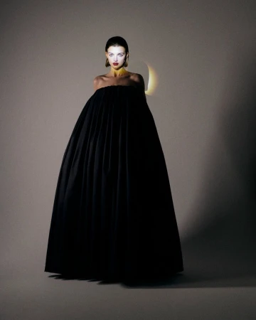 Model wearing an expansive strapless black pleated ball gown standing against a neutral studio backdrop with focused backlighting.
