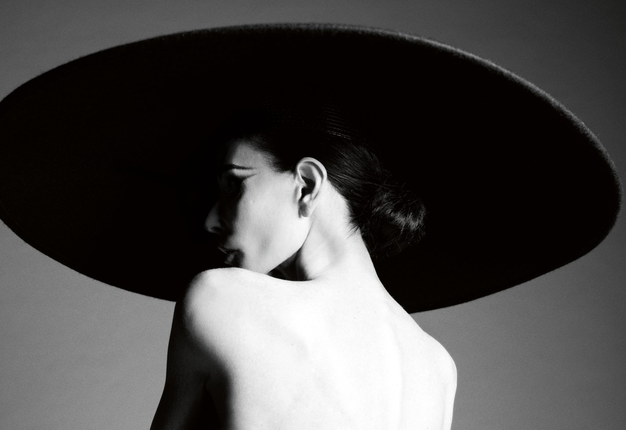 Monochrome profile portrait of Demi Moore wearing an oversized black wide-brim hat with bare shoulders against a grey studio backdrop.