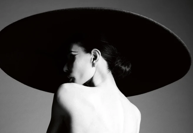 Monochrome profile portrait of Demi Moore wearing an oversized black wide-brim hat with bare shoulders against a grey studio backdrop.