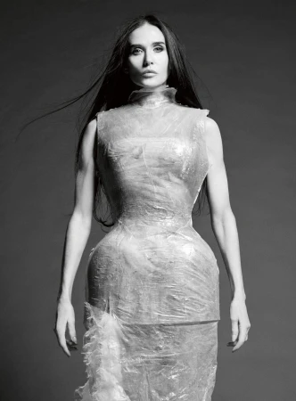 Demi Moore posing in a translucent sculptural plastic wrap dress against a grey studio backdrop in a black and white portrait.