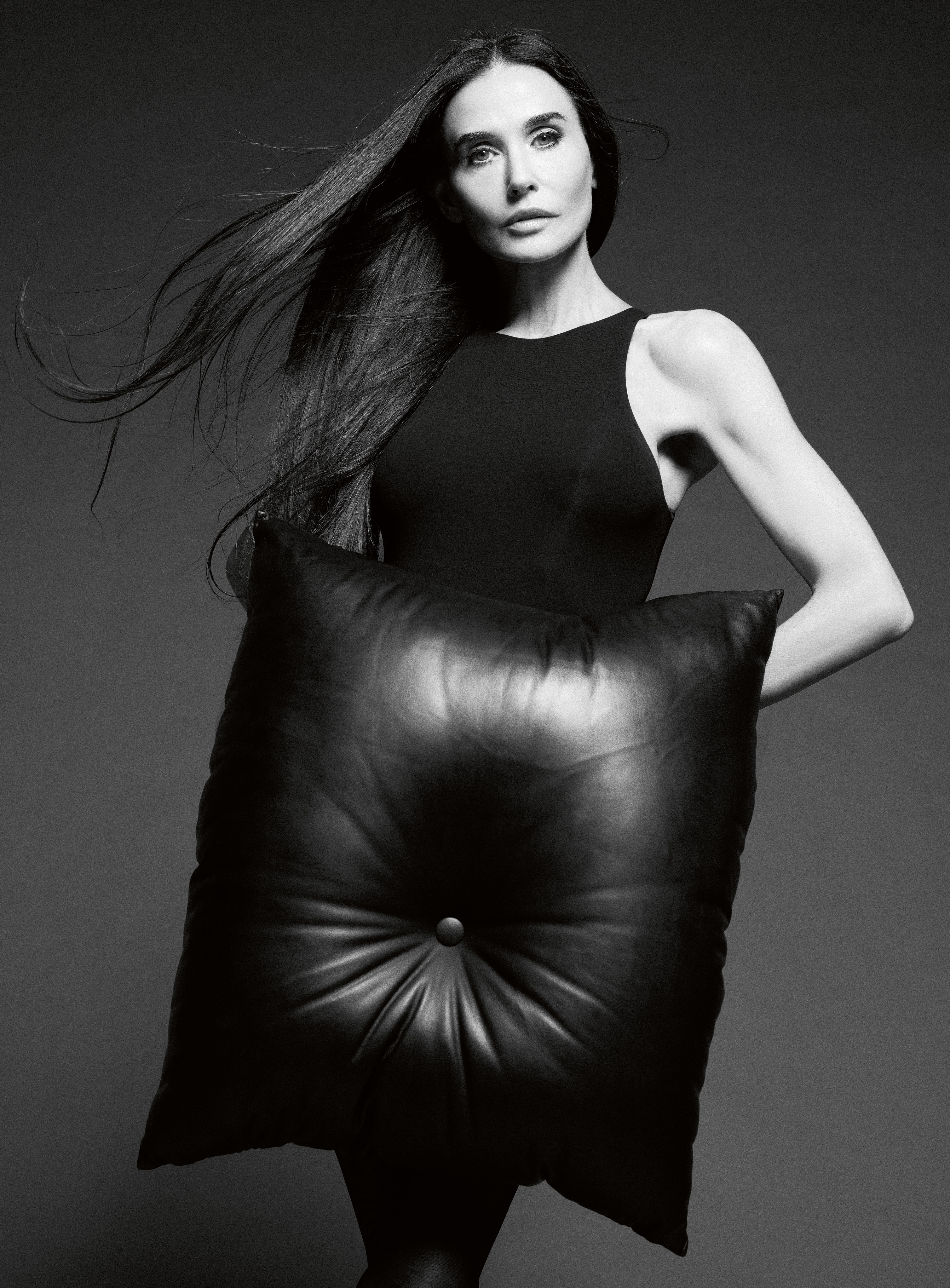 Demi Moore posing with an oversized tufted black leather pillow in a black and white studio portrait.