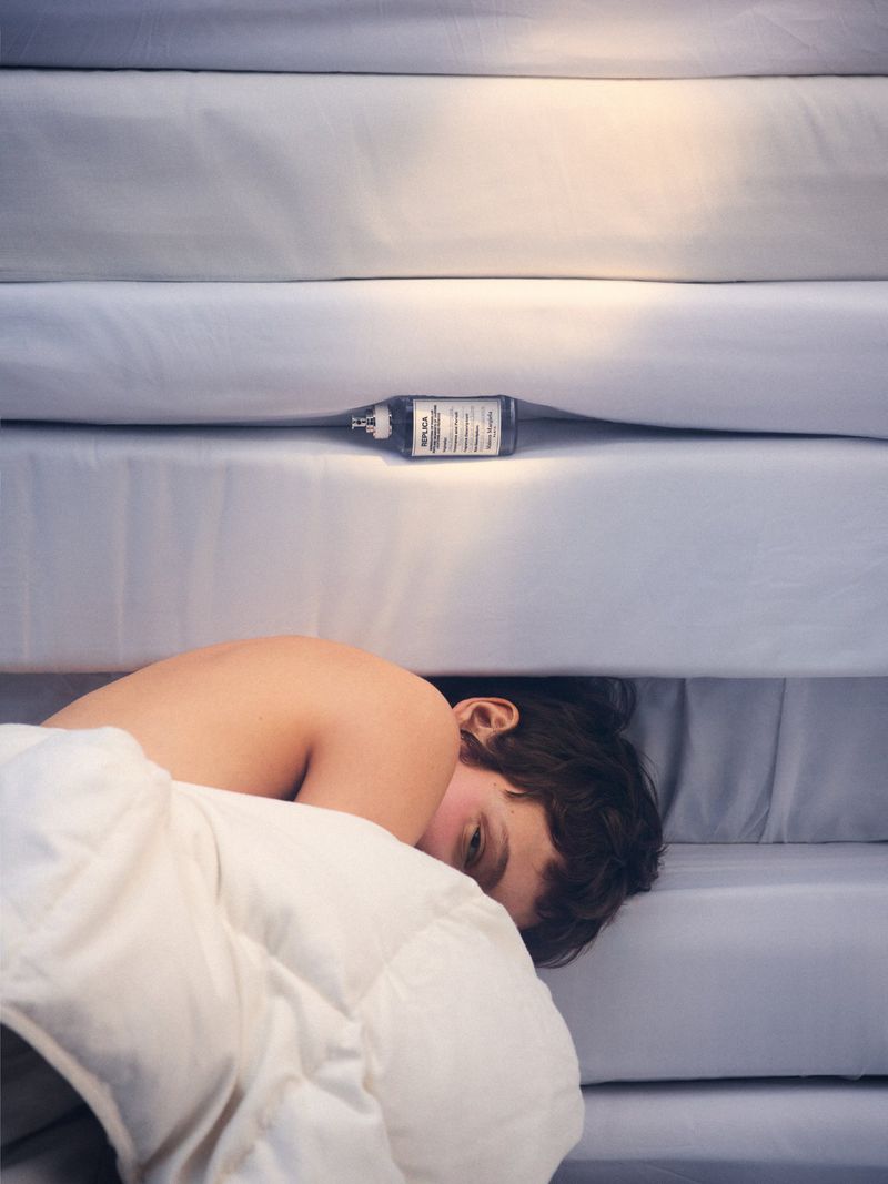 A person with dark hair lies nestled in white bedding with a small perfume bottle placed neatly on the crisp, layered mattress folds above.
