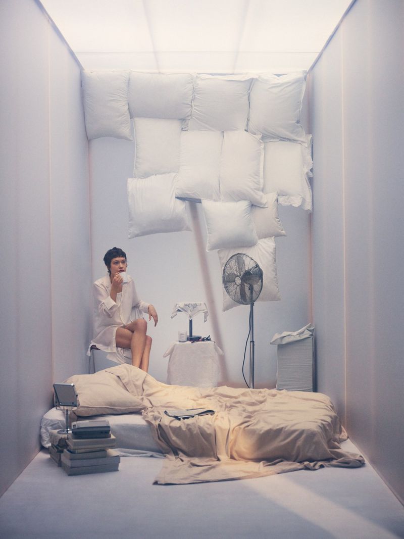 Woman in a white shirt seated in a minimalist room with an unmade floor mattress, fan, and pillows arranged on the back wall.