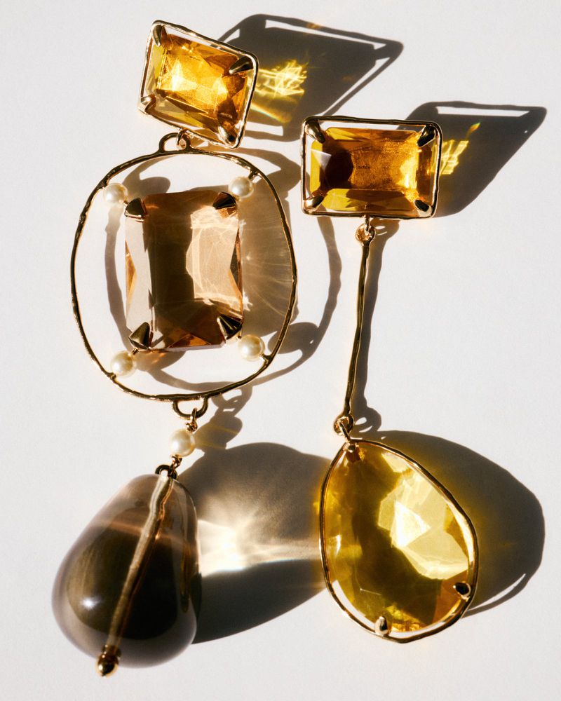 Two mismatched Givenchy earrings with amber-colored crystals and small pearls, casting sharp shadows on a plain white surface.