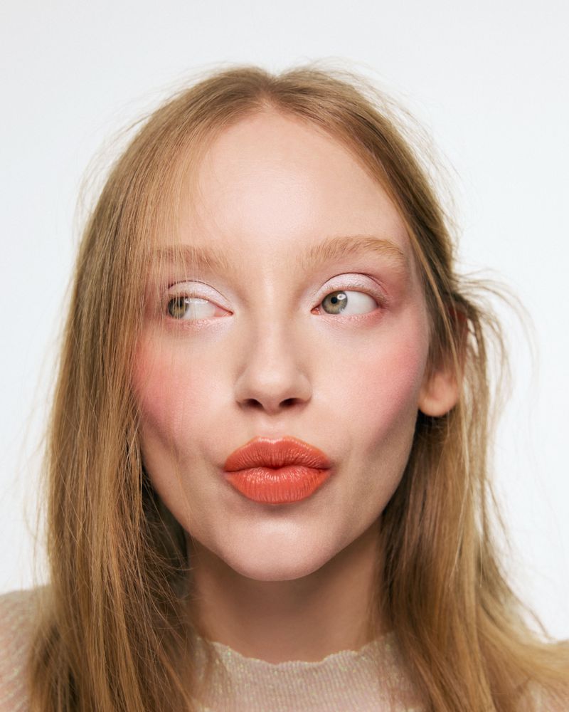Close-up portrait of a fair-skinned woman with long blonde hair, pouting her bright orange-lipstick lips against a stark white background.
