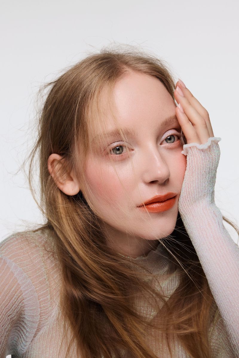 Close-up portrait of a woman wearing warm orange lipstick and a sheer textured knit top against a plain white studio backdrop.
