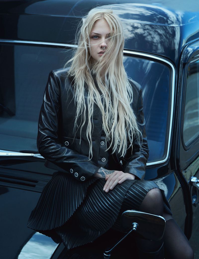 A fair-skinned woman with platinum blonde hair poses, seated on a black vintage car, wearing a black leather jacket and pleated skirt.