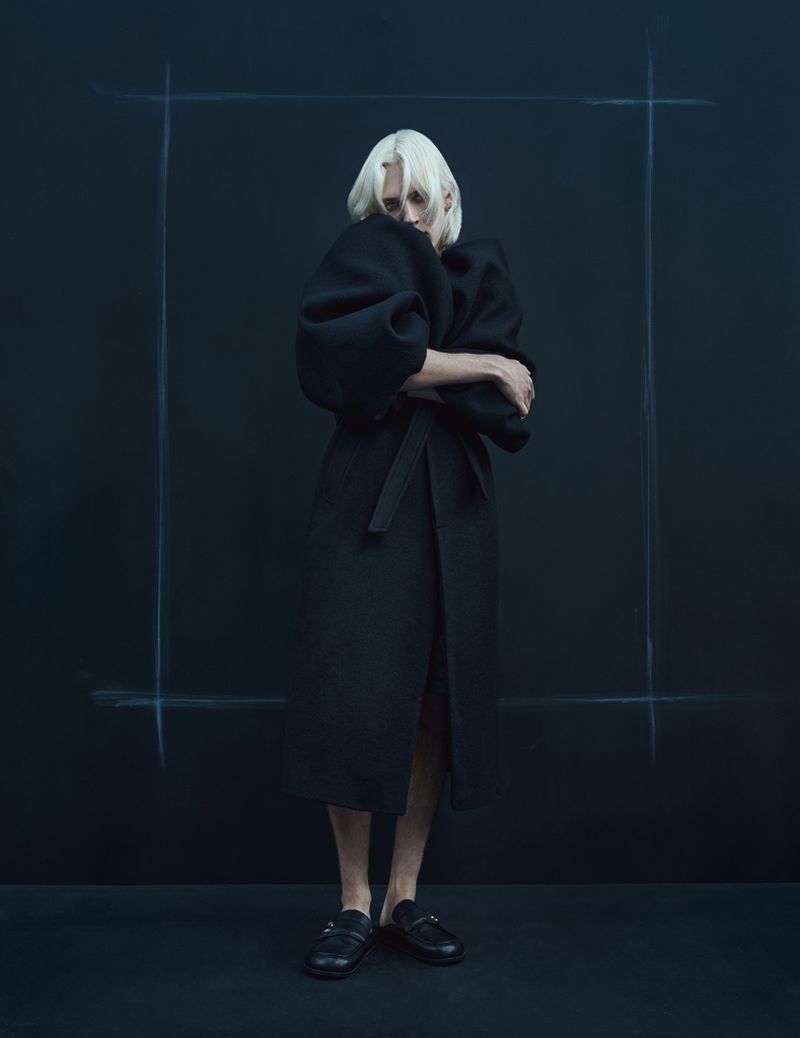 Woman with platinum blonde hair in a long black coat with puffy sleeves poses against a dark background with a simple frame outline.