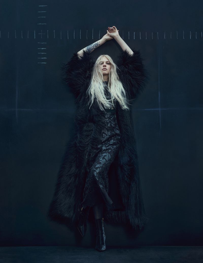 A full-length portrait of a blonde woman in a long black gown and feathered coat, standing in front of a dark wall with white markings.