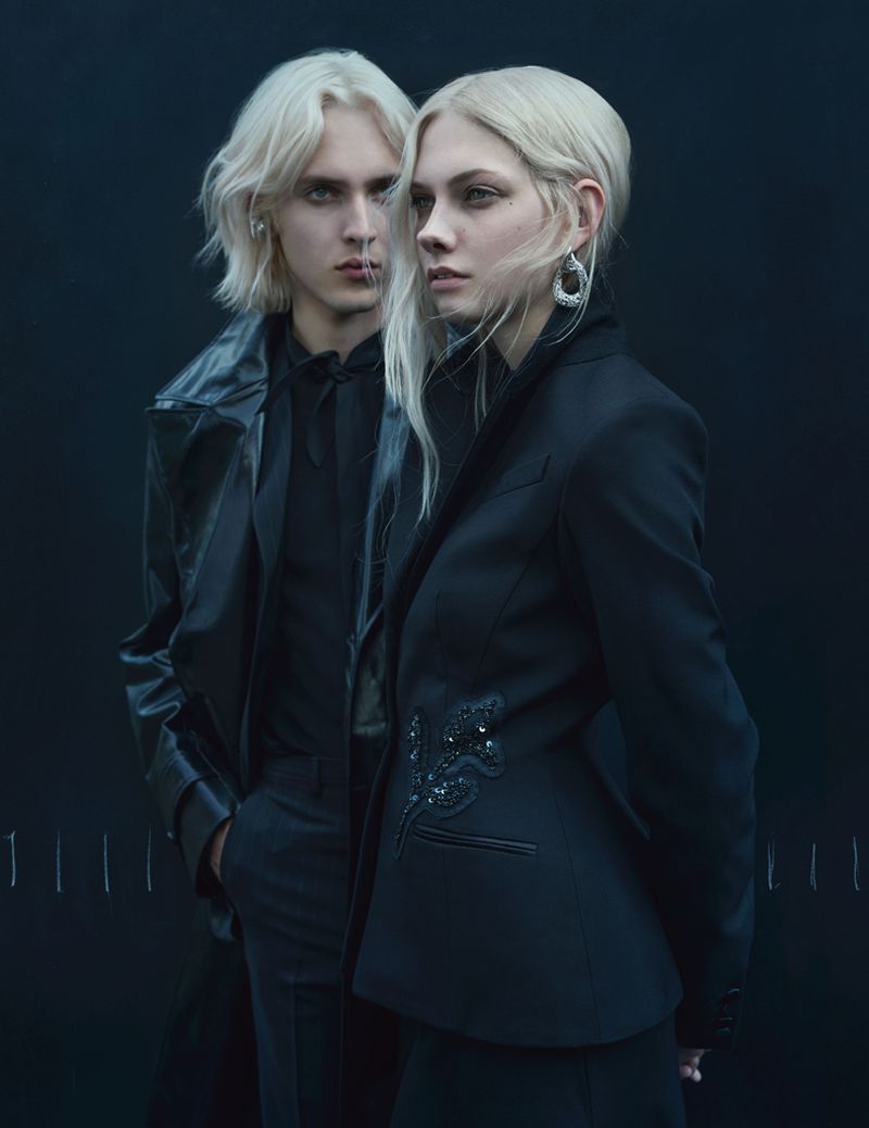 Two fair-skinned people with fair hair pose in black tailored clothing against a dark backdrop. The composition is studio-lit, showcasing fashion.