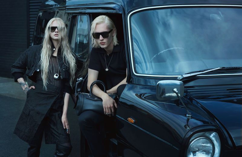 Two models with blonde hair, sunglasses, and dark clothing pose next to a black taxicab in an urban setting, creating a fashion-editorial look.