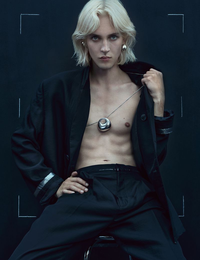 Portrait of a blonde adult male with bare torso, wearing jewelry, a blazer, and trousers in a studio setting against a dark background.