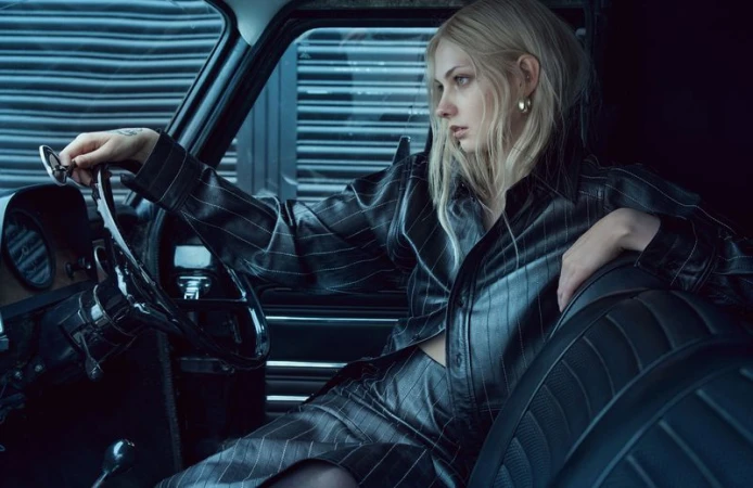 A blonde woman sits in the driver's seat of a vintage car wearing a pinstripe suit and gold hoop earrings, holding glasses.