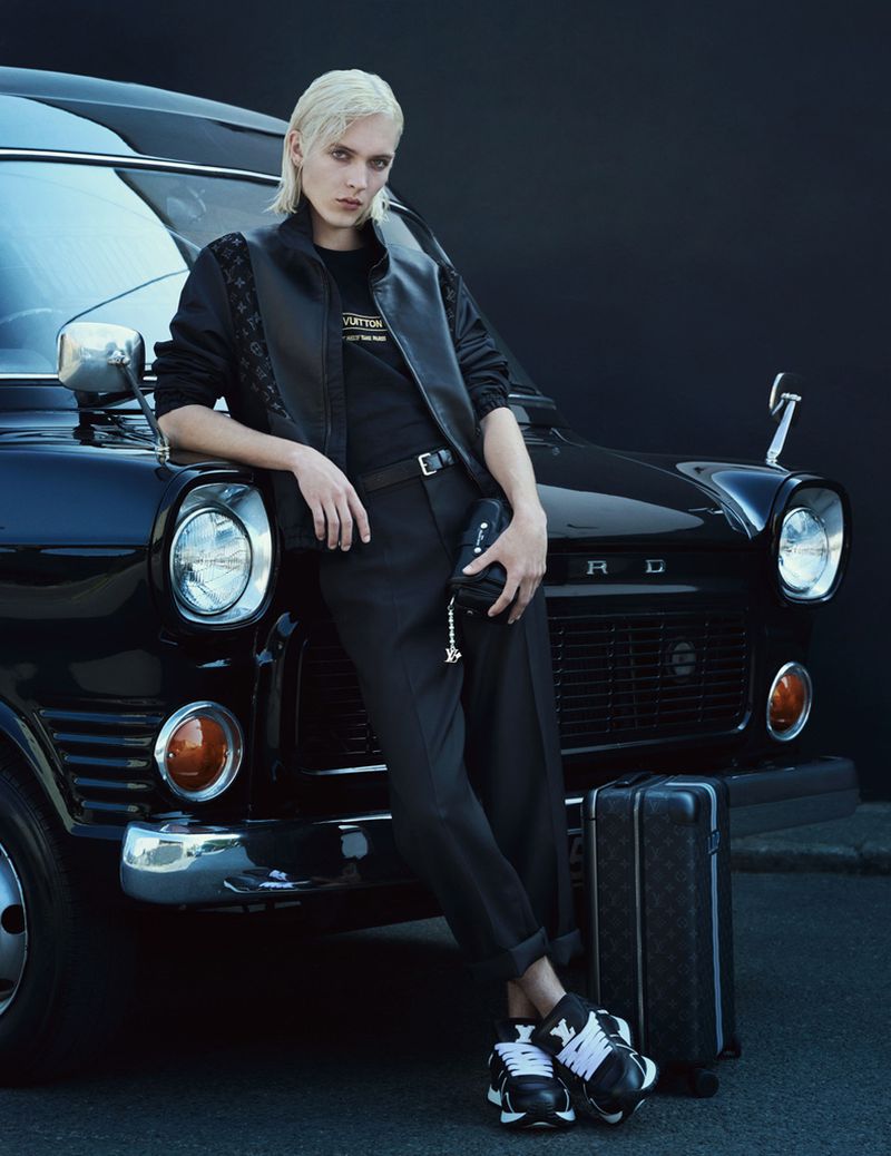 An androgynous model poses leaning against a vintage black Ford van in a fashion shot. Suitcase, all-black outfit, black shoes.