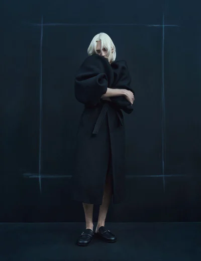 Model with platinum blonde hair wearing a long black coat against a dark wall with chalk framing lines.