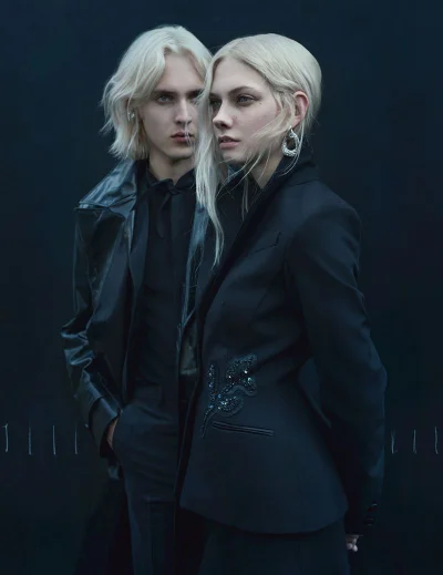 Delice Rimmer and Dylan Moffett posing in black tailored outerwear with platinum blonde hair against a dark studio backdrop.