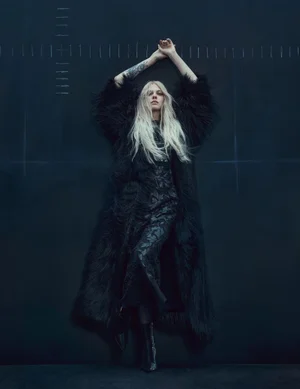 Model with blonde hair posing with arms raised in a long black shaggy coat and dark patterned dress against a dark studio backdrop.