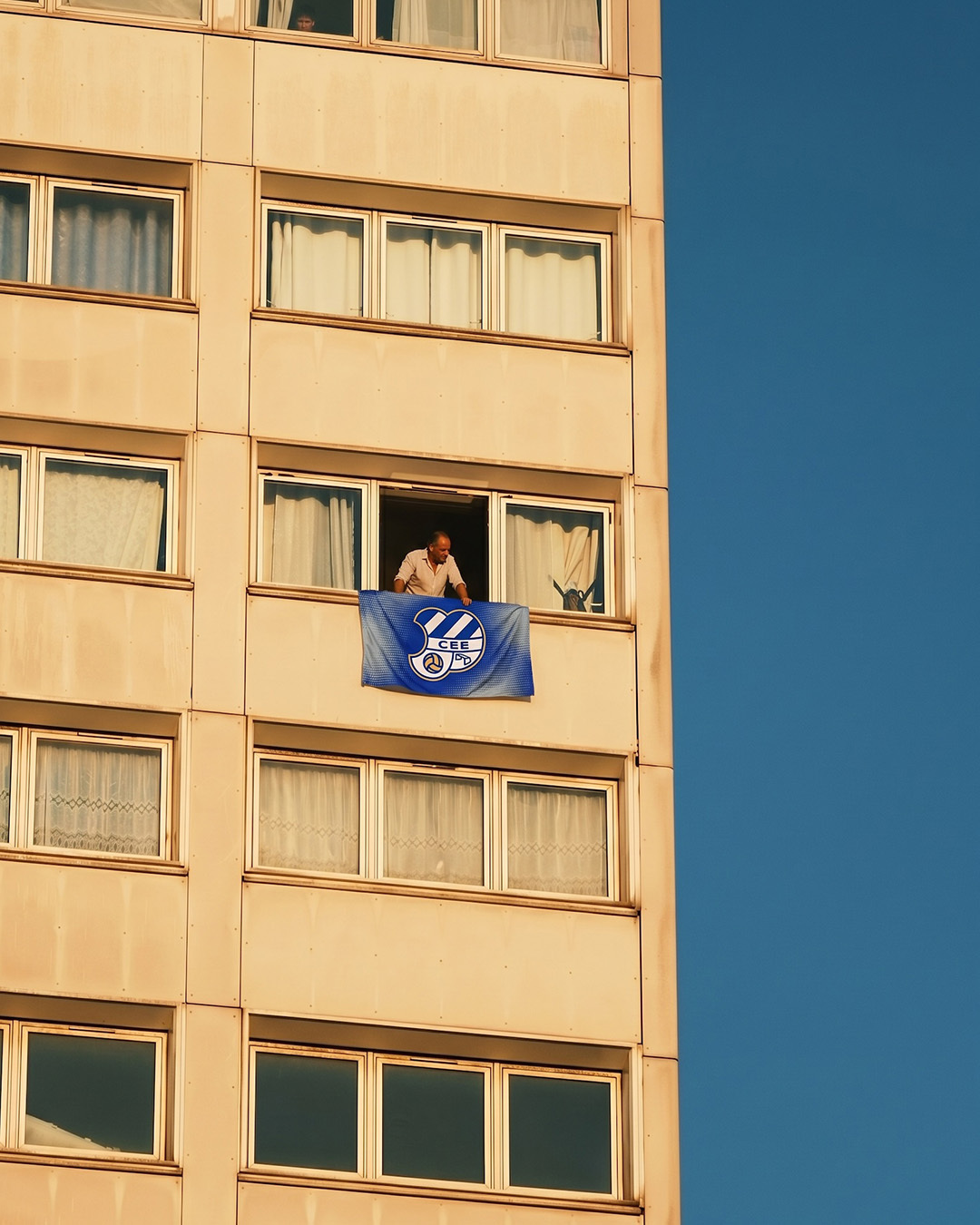 A man leans out from a high-rise window, holding a blue CE Europa sports flag against a clear deep blue sky.