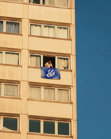 A man leans out from a high-rise window, holding a blue CE Europa sports flag against a clear deep blue sky.