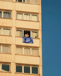 Man leaning out of a multi-story apartment window with a blue CE Europa flag hung over the ledge.
