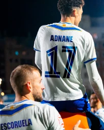 Soccer player seen from behind in a white CE Europa jersey numbered 17 Adnane celebrating with teammates under stadium lights.