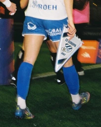 Soccer player from behind holding a CE Europa triangular pennant, wearing blue athletic shorts, high blue socks, and cleats on a pitch.