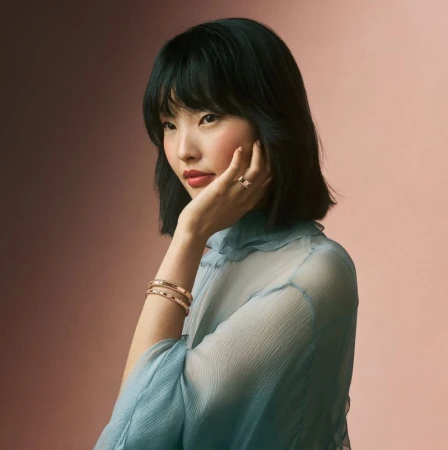 East Asian woman with a black bob haircut wearing a blue sheer ruffled blouse, showcasing gold rings and bracelets against a soft pink background.