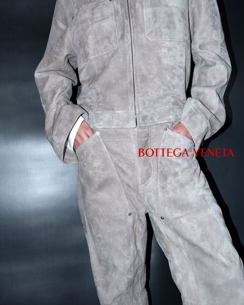 A person wearing a grey suede workwear-style jacket and matching trousers with hands tucked in pockets, against a dark backdrop.